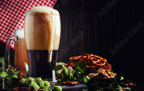 Dark German beer is poured into a glass, fresh green hops and bowls with salty snacks and nuts, autumn beer festival concept, dark background, selective focus photo