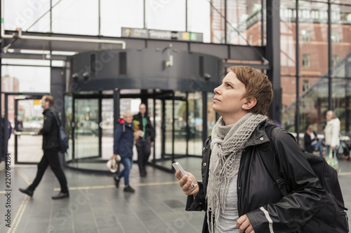 Thoughtful woman holding smart phone while looking up by building in city photo