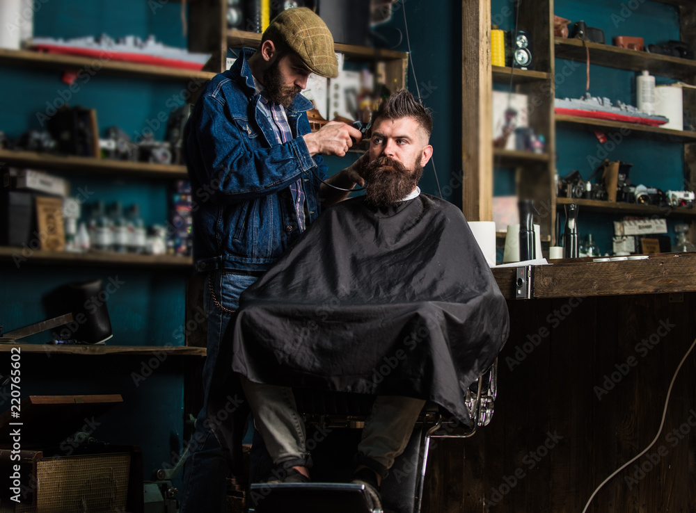 Barber with clipper trimming hair on temple of bearded client. Hipster client getting haircut. Barber with hair clipper work on hairstyle for hipster, barbershop background. Hipster lifestyle concept