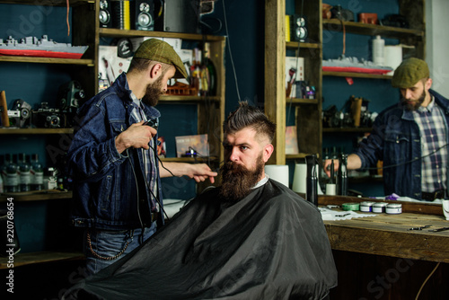 Barber preparing hair clipper for bearded man, barbershop background. Barber with clipper and brutal bearded client. Hipster client covered with cape getting haircut. Hipster lifestyle concept