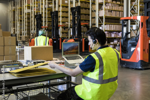 Rear view of confident young male customer service representative looking at package while sitting with laptop in wareho photo