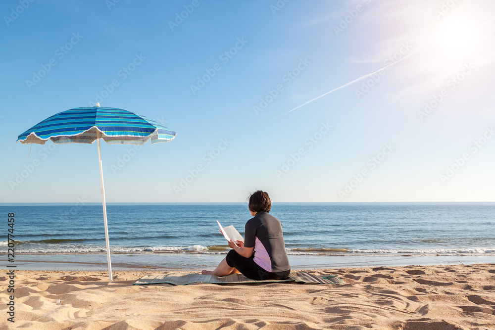 A girl on the beach, near the sea reading a book. Summer on the sand.