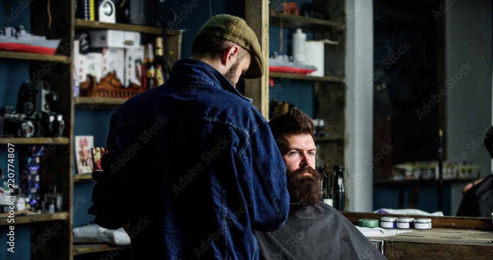 Hipster with beard covered with cape serving by professional barber in stylish barbershop. Barber busy with grooming beard of hipster client, beauty supplies on background. Grooming concept