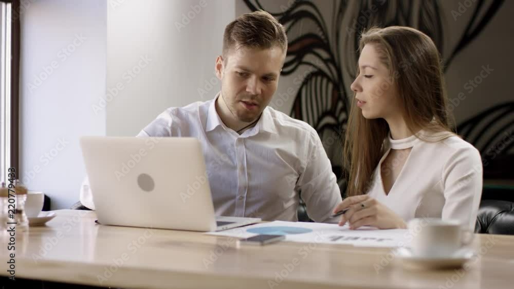 Two young people work with a laptop in a cafe. They are discussing and preparing the contract. It is a business meeting. Man and woman are sitting in the cafe at the table during break. Couple are Two young people work with a laptop in a cafe. They are discussing and preparing the contract. It is a business meeting. Man and woman are sitting in the cafe at the table during break. Couple are