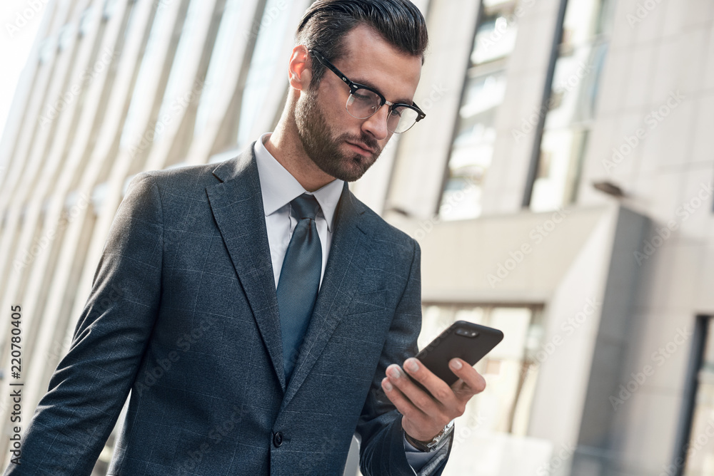 Good looking young man in full suit using his smart phone while standing outdoors Good looking young man in full suit using his smart phone while standing outdoors