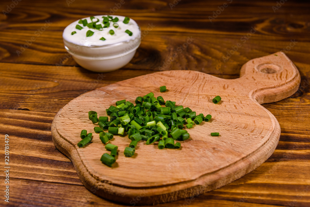 Glass bowl with sour cream and cutting board with chopped green onion on wooden table