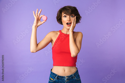 Image of european young woman 20s in casual wear smiling and holding macaron biscuit, isolated over violet background