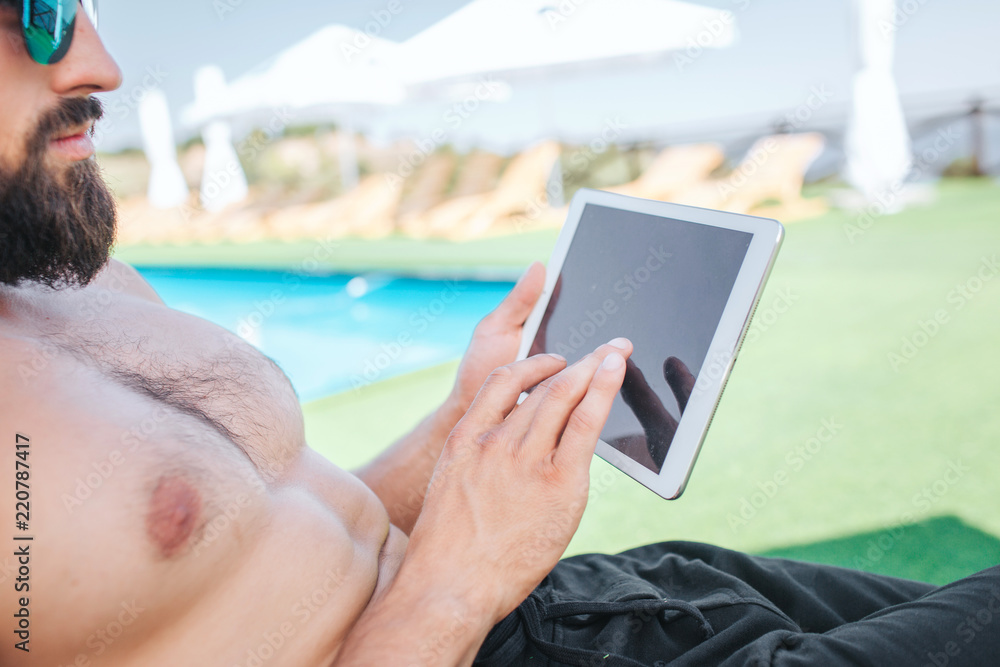 Cut view of bearded guy sits on sunbed and wears glasses. He holds white tablet and looks at it. He works. Guy is concentrated.