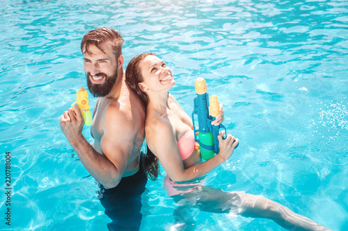 Funny couple stands in swimming pool. They pose and smile. Girl looks up. They hold water guns in hands. They are ready to shoot.