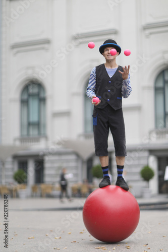 The clown juggles with pink balls, standing on a big red ball in the street of a European city