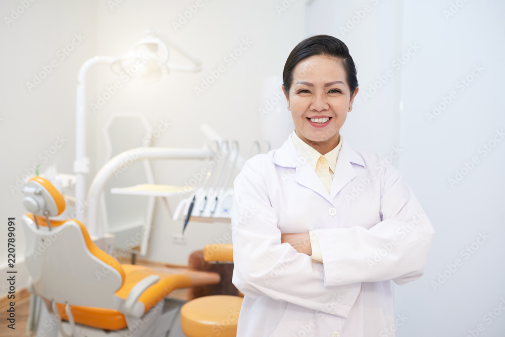 Adult smiling Asian woman in medical gown holding hands crossed and smiling at camera in office Adult smiling Asian woman in medical gown holding hands crossed and smiling at camera in office