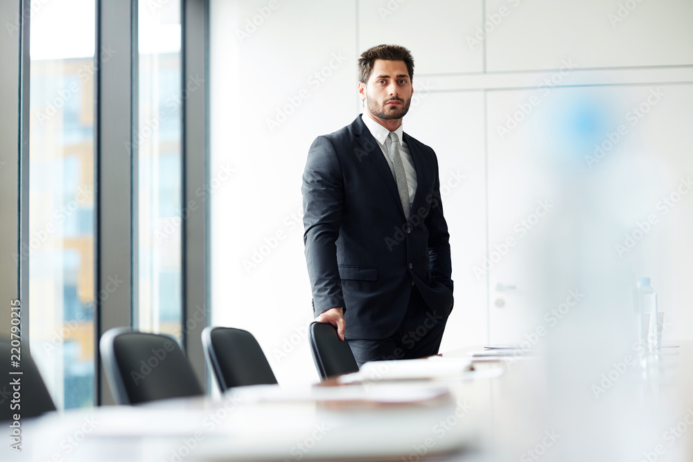 Portrait of successful businessman standing by table in conference room and looking at camera, copy space Portrait of successful businessman standing by table in conference room and looking at camera, copy space