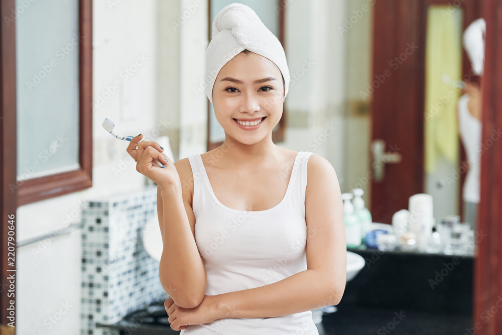 Young cheerful Asian woman with towel on head holding toothbrush standing in bathroom smiling at camera Young cheerful Asian woman with towel on head holding toothbrush standing in bathroom smiling at camera