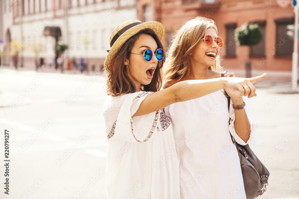 Naklejka premium Fashion portrait of two young stylish hippie brunette and blond women models in summer sunny day in white hipster clothes posing on the street background. Pointing on shop sales