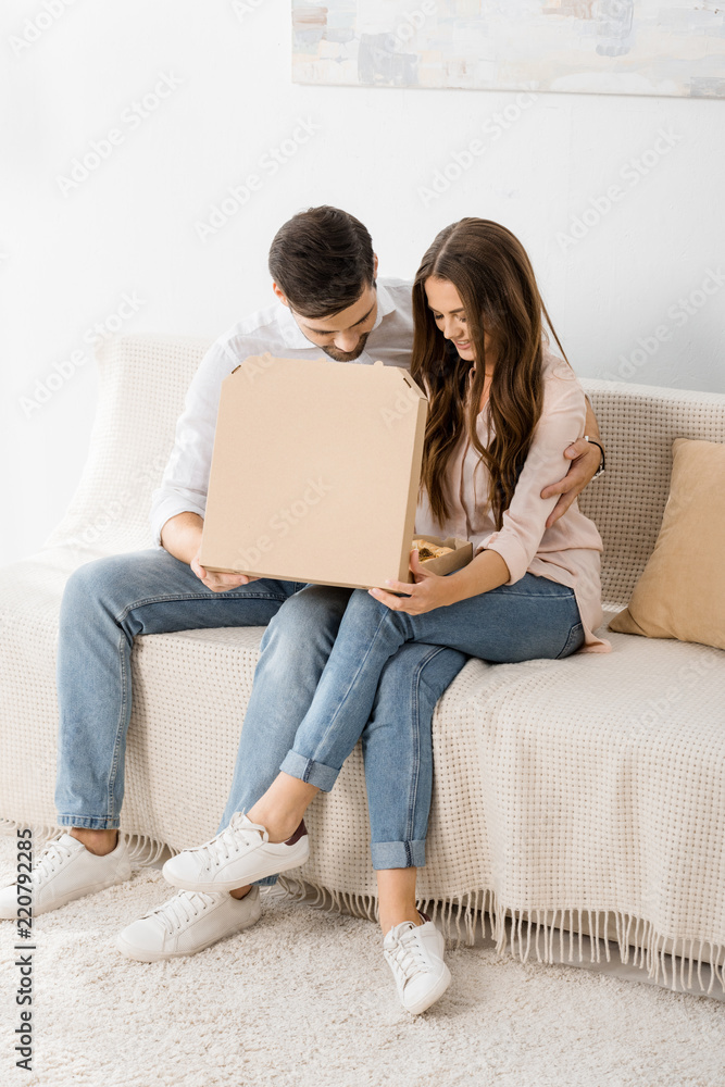 smiling couple with pizza in paper box sitting on sofa at home smiling couple with pizza in paper box sitting on sofa at home