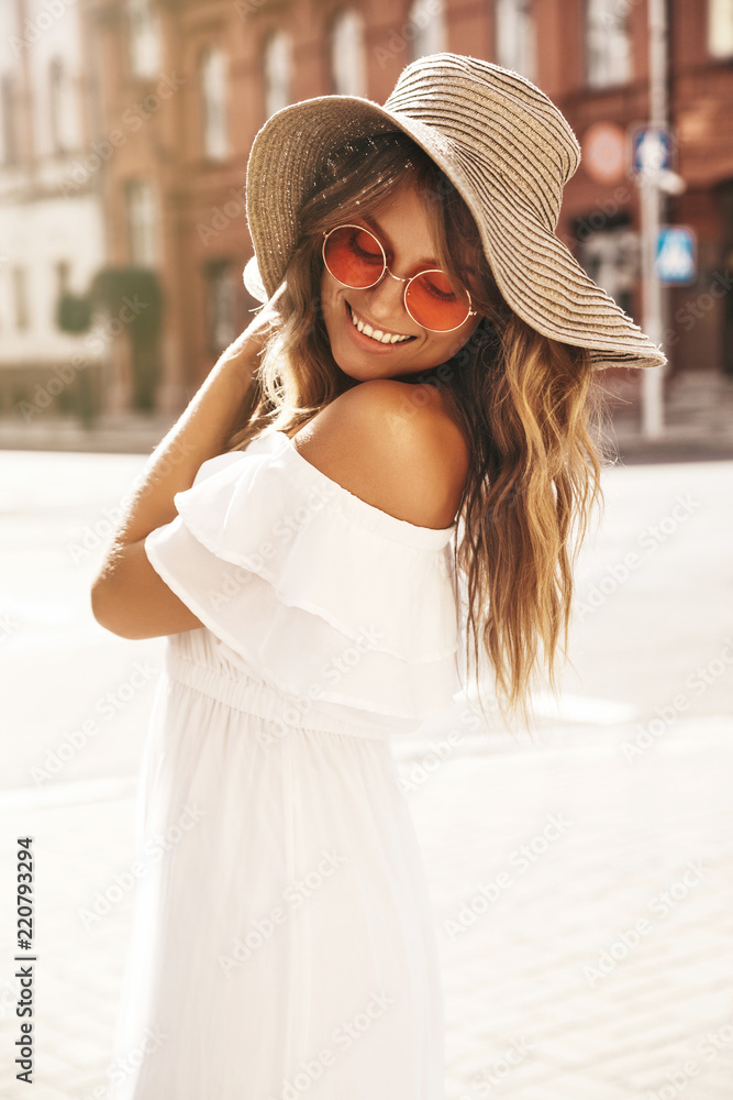 Portrait of beautiful smiling cute blond teenager model with no makeup in summer hipster white dress and big beach hat posing on the street background