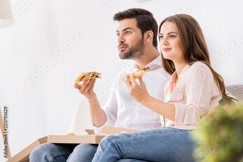 portrait of young couple eating pizza while watching tv at home