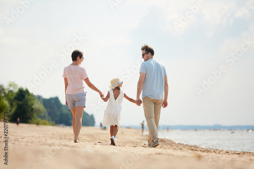 Back view portrait of happy family holding hands while enjoying walk on beach during Summer vacation