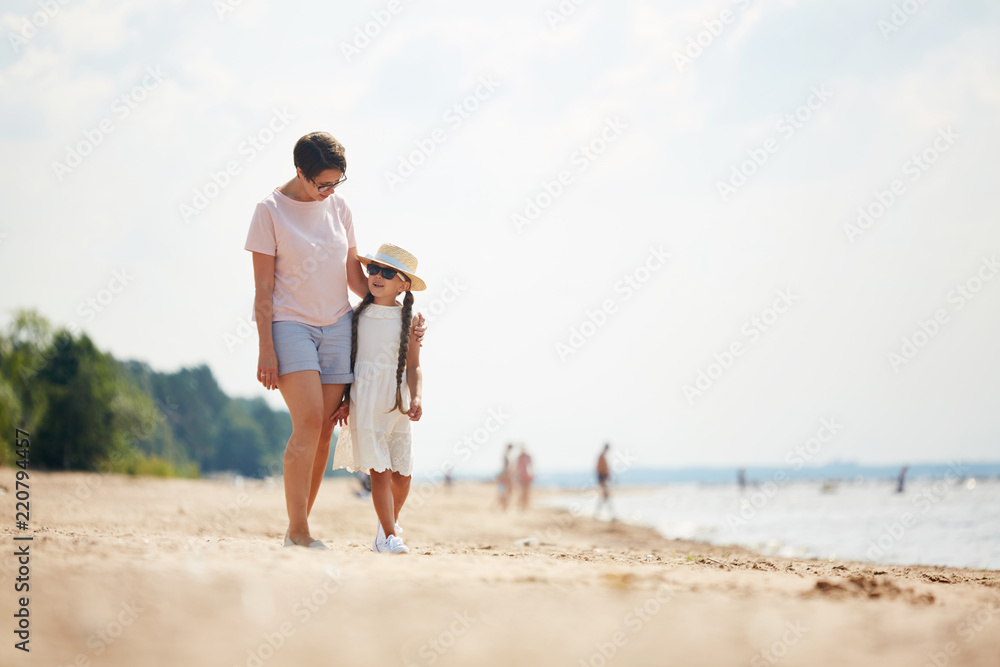 Full length portrait of loving modern mom embracing daughter while enjoying walk along beach during Summer vacation, copy space Full length portrait of loving modern mom embracing daughter while enjoying walk along beach during Summer vacation, copy space