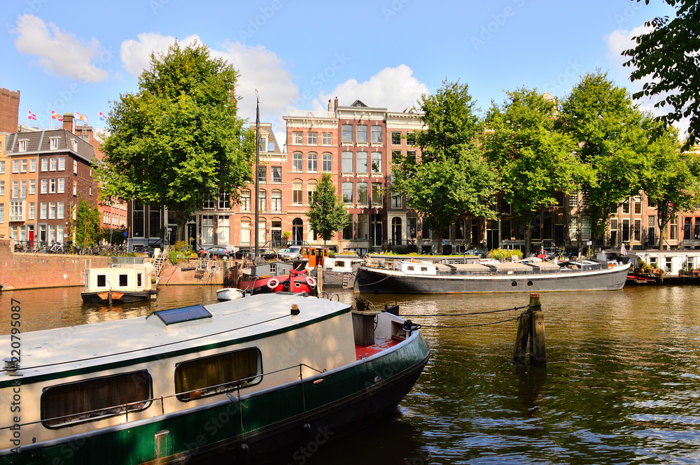 Kanał w Amsterdamie, barka, zabytkowe kamienice i zielone drzewa.