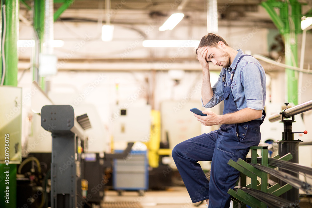 Embarrassed young engineer of factory reading notification in smartphone and touching his forehead Embarrassed young engineer of factory reading notification in smartphone and touching his forehead