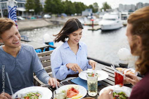 Happy young friends enjoying lunch by waterside in outdoor cafe and having talk