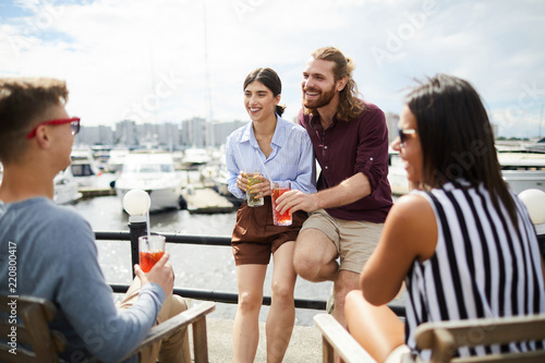Happy young man and woman with drinks having talk to their friends by waterside on summer day © pressmaster