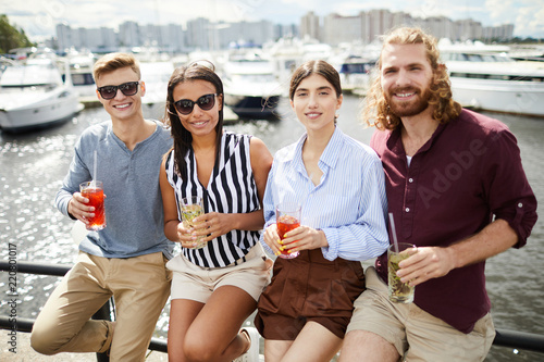 Row of four young friends in casualwear holding drinks while having rest by waterside on summer day