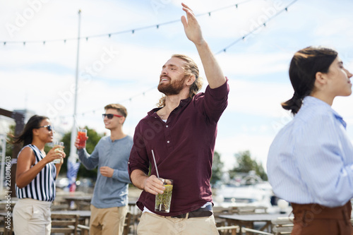 Young ecstatic man with glass of drink dancing outdoors among his friends