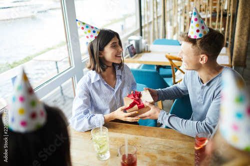 Happy guy congratulating his girlfriend on birthday while sitting in cafe with friends