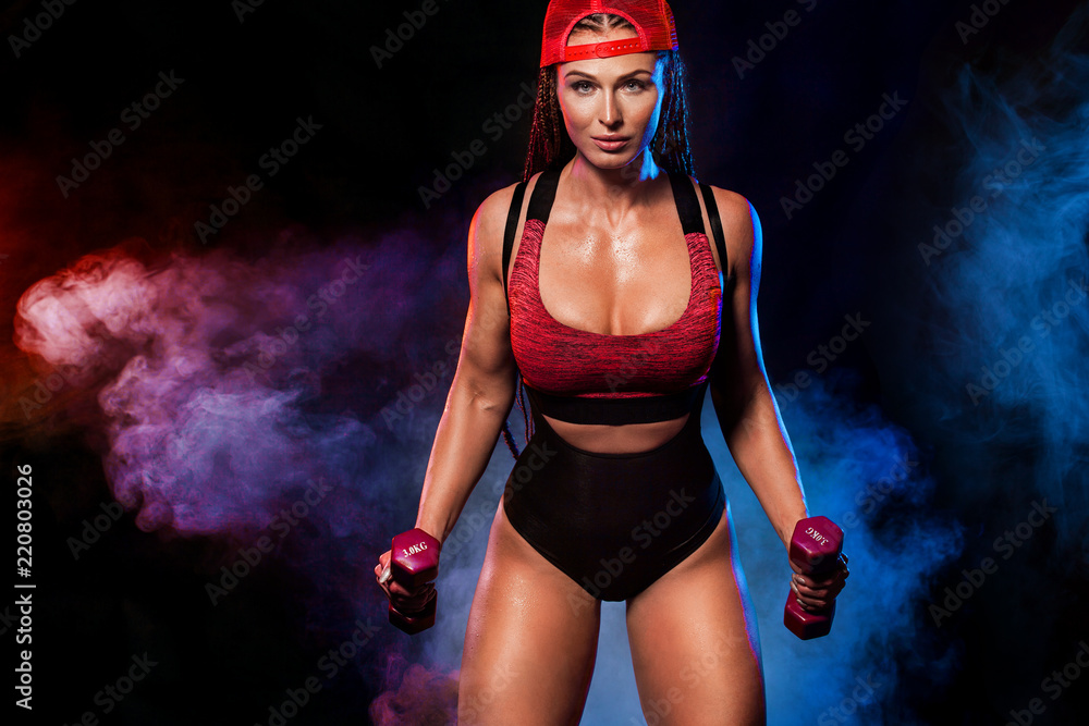 Obraz premium Attractive young fitness woman athlete and bodybuilder holding dumbbell on black background with color smoke. Copy space for fitness nutrition ads.