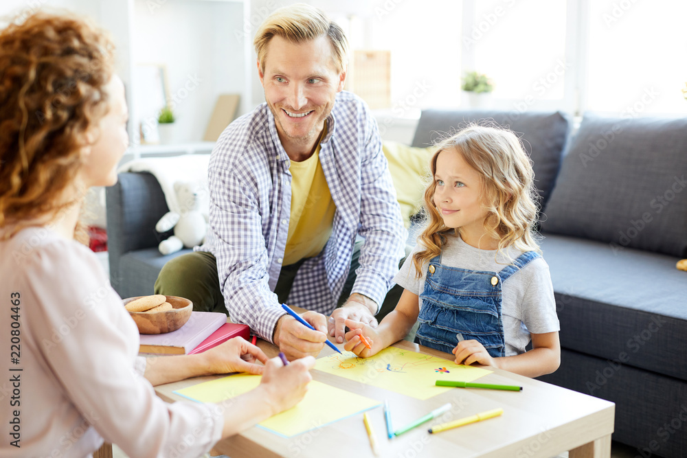 Happy father helping little daughter with drawing while talking to young mother