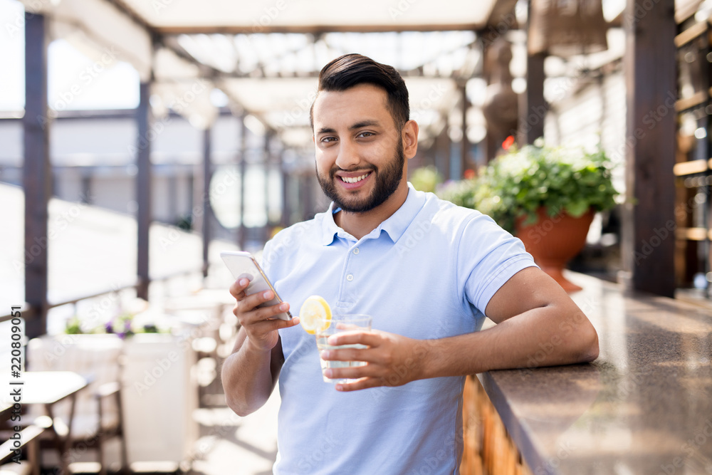 Waist up portrait of handsome Middle-Eastern man looking at camera and smiling while using smartphone leaning on bar counter in outdoor terrace