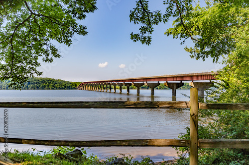 Slika na platnu View over split rail fence of bridge over the Tennessee river on the Natchez Trace Parkway in Mississippi, USA, with blue sky and framed on both sides by the spring green of hardwood branches