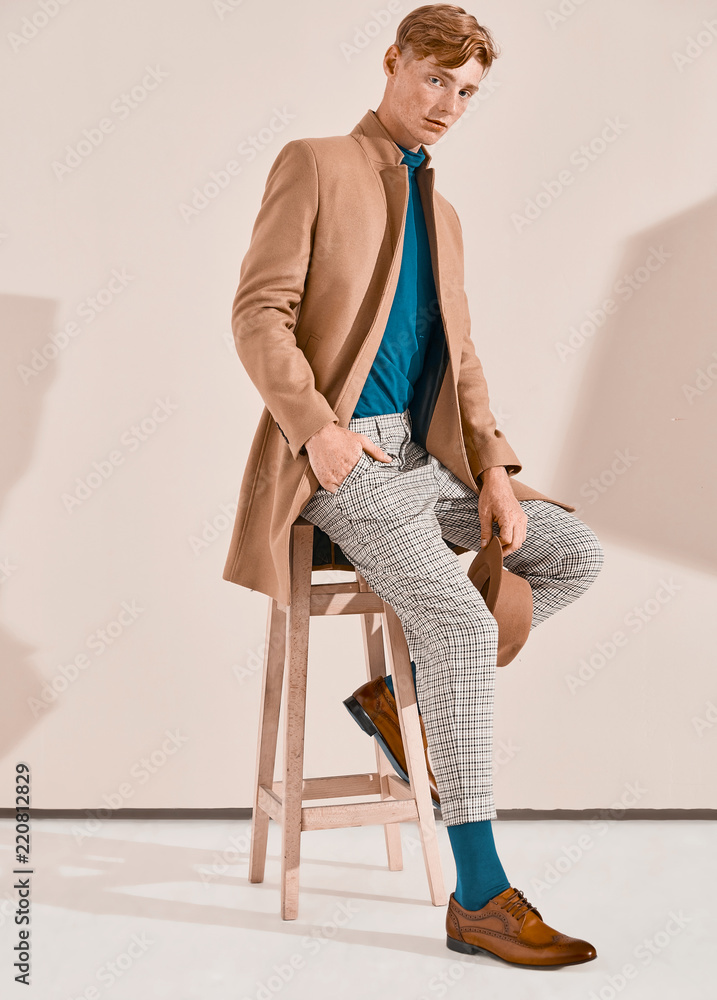 Young man with red hair and freckles posing in brown coat and hat, plaid  pants, turquoise sweater and socks, brown shoes, sitting on chair. Pink  background Stock Photo | Adobe Stock