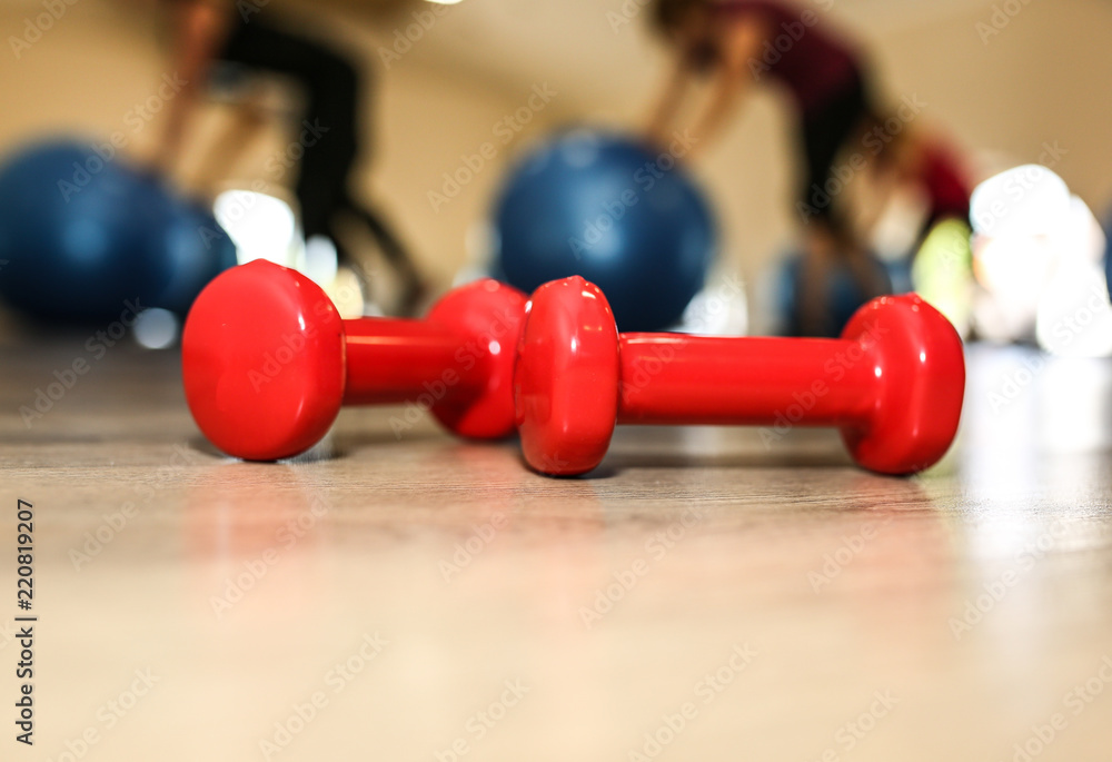 Fototapeta premium Small red dumbbells on the wooden floor. Fitness gears in the gym with girls doing exercises with balls on the background. Sport and healthy lifestyle concept. Women are getting fit.