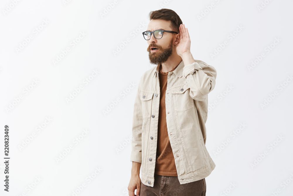 Studio shot of intense intrigued adult husband with long beard and cool hairstyle turning to camera with ear and holding palm near it while eavesdropping or overhearing interesting conversation