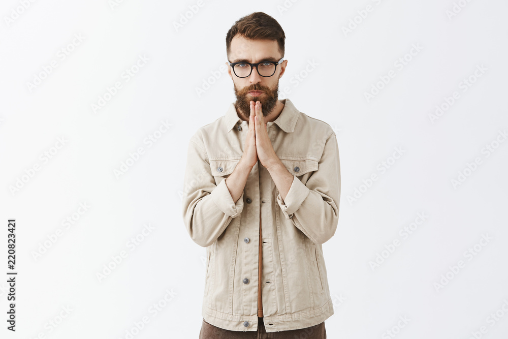 Indoor shot of gloomy unhappy and sad adult man with long beard in beige outdoor jacket holding hands in pray standing with serious unemotional expression begging for money or praying Indoor shot of gloomy unhappy and sad adult man with long beard in beige outdoor jacket holding hands in pray standing with serious unemotional expression begging for money or praying