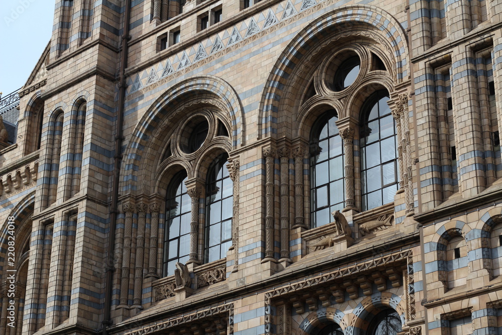  Large gothic windows of the London Museum of Natural Sciences, placed on a light and blue brick wall with the presence of some gargoyles