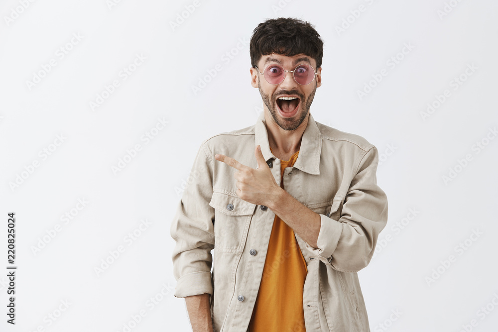 Studio shot of excited and joyful party guy in stylish sunglasses and trendy jacket dropping jaw from amazement and astonishment pointing at upper left corner being impressed over gray background Studio shot of excited and joyful party guy in stylish sunglasses and trendy jacket dropping jaw from amazement and astonishment pointing at upper left corner being impressed over gray background
