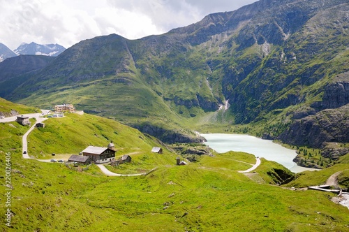 A blue mountain lake between the green slopes of the mountains. Several small houses and hotel buildings. Beautiful curving mountain road, Grossglockner Hochalpenstrasse, Austria. Summer day.