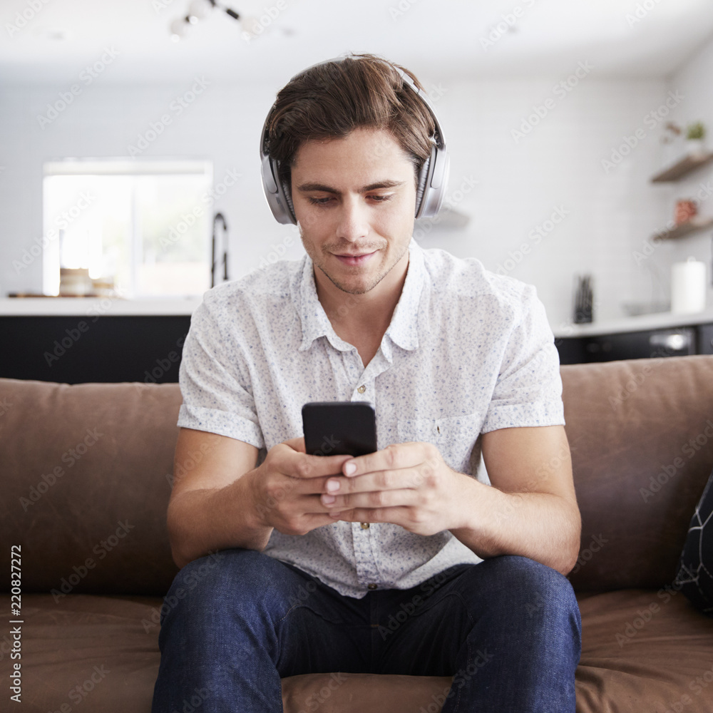 Man Wearing Wireless Headphones Sitting On Sofa At Home Streaming From Mobile Phone Man Wearing Wireless Headphones Sitting On Sofa At Home Streaming From Mobile Phone