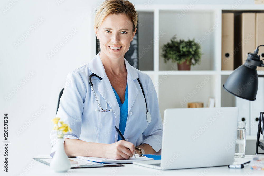 adult female doctor with stethoscope over neck writing in clipboard at table with laptop in office