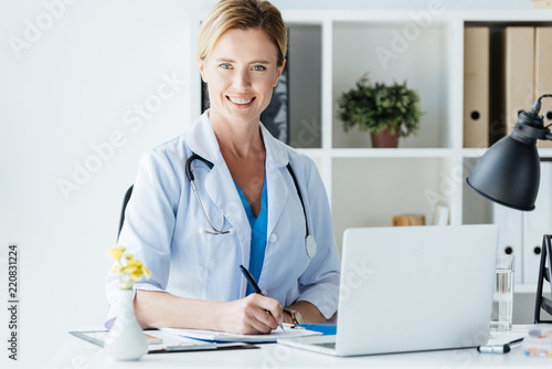 adult female doctor with stethoscope over neck writing in clipboard at table with laptop in office