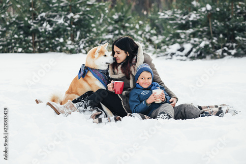 Little child and mom plays with funny Akita-inu dog in a winter park. Christmas happy family,mother and son walking with dog lying on snow in winter day. Drinking hot coffee or tea on snowy winter