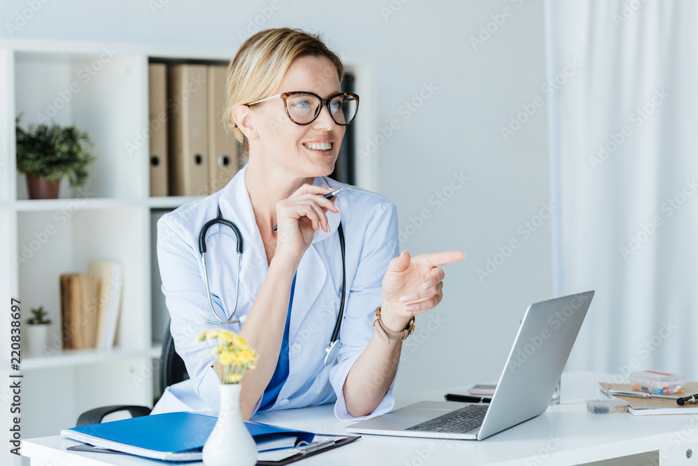 smiling female doctor in eyeglasses pointing by finger and talking at table with laptop in office smiling female doctor in eyeglasses pointing by finger and talking at table with laptop in office