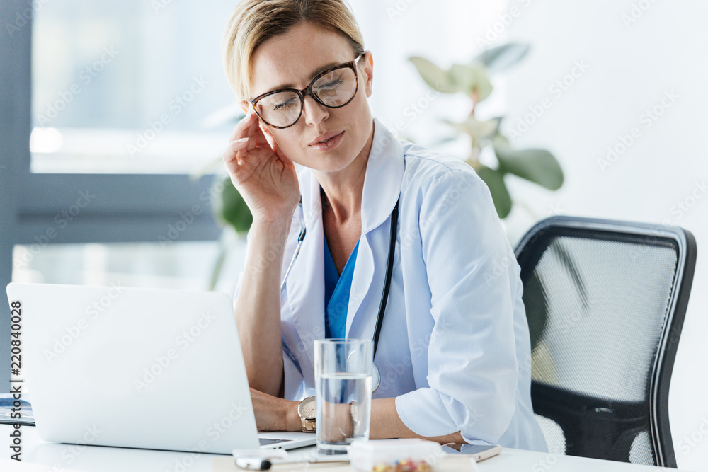 exhausted female doctor in eyeglasses having headache at table in office exhausted female doctor in eyeglasses having headache at table in office