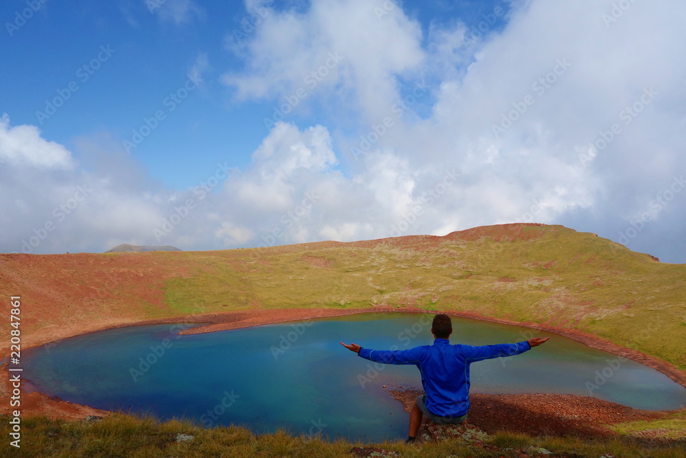 Young man sitting at the edge of Azhdahak volcano with a turquoise lake hidden inside in Geghama mountains, Armenia