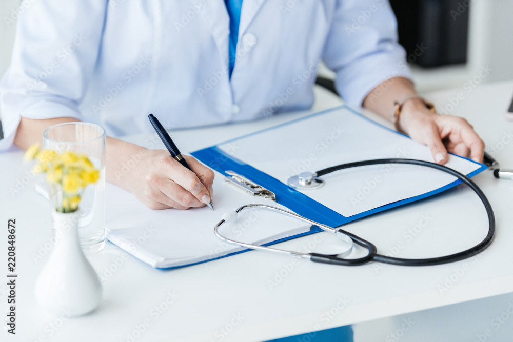 Fototapeta premium cropped image of female doctor writing in clipboard at table with stethoscope in office