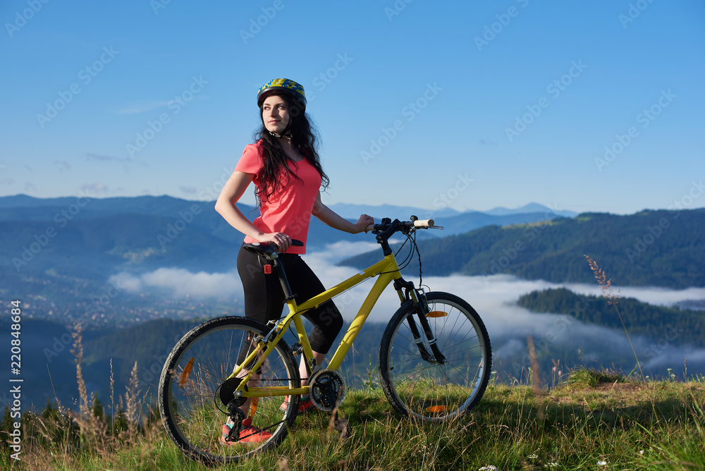 Obraz premium Attractive woman cyclist with yellow bike on a rural trail in the mountains, wearing helmet and red red t-shirt, enjoying morning haze in valley, forests on blurred background. Outdoor sport activity